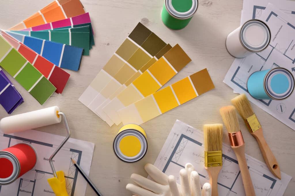 Palette de couleurs, pots de peinture (rouge, jaune, bleu ciel, vert, blanc), rouleau, pinceaux et plans d'étage pour projet de rénovation intérieure.